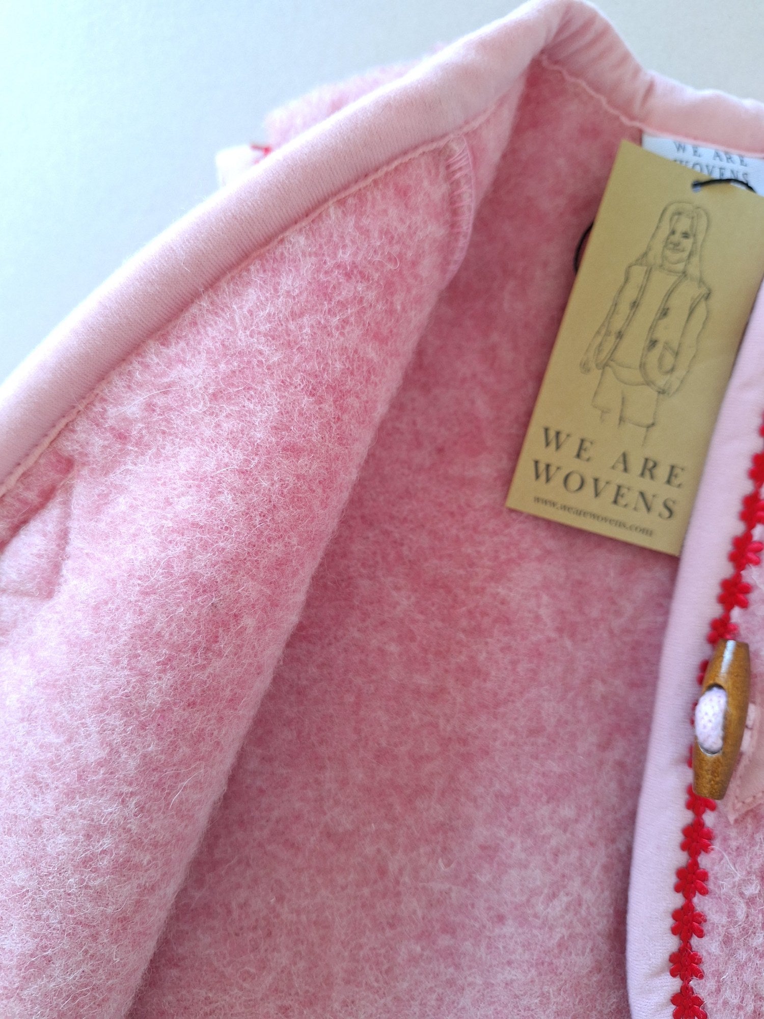 Close-up of inside of pink wool fabric of kids bodywarmer with a 'WE ARE WOVENS' tag on a cream background