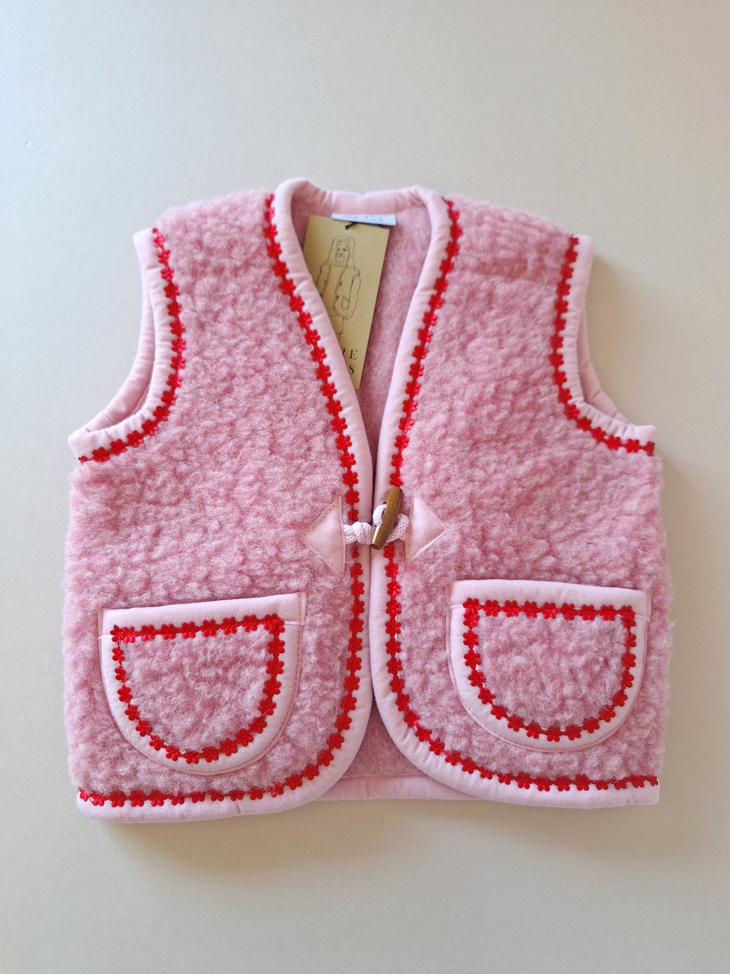 Pink children's wool bodywarmer with red flower trim on a cream background