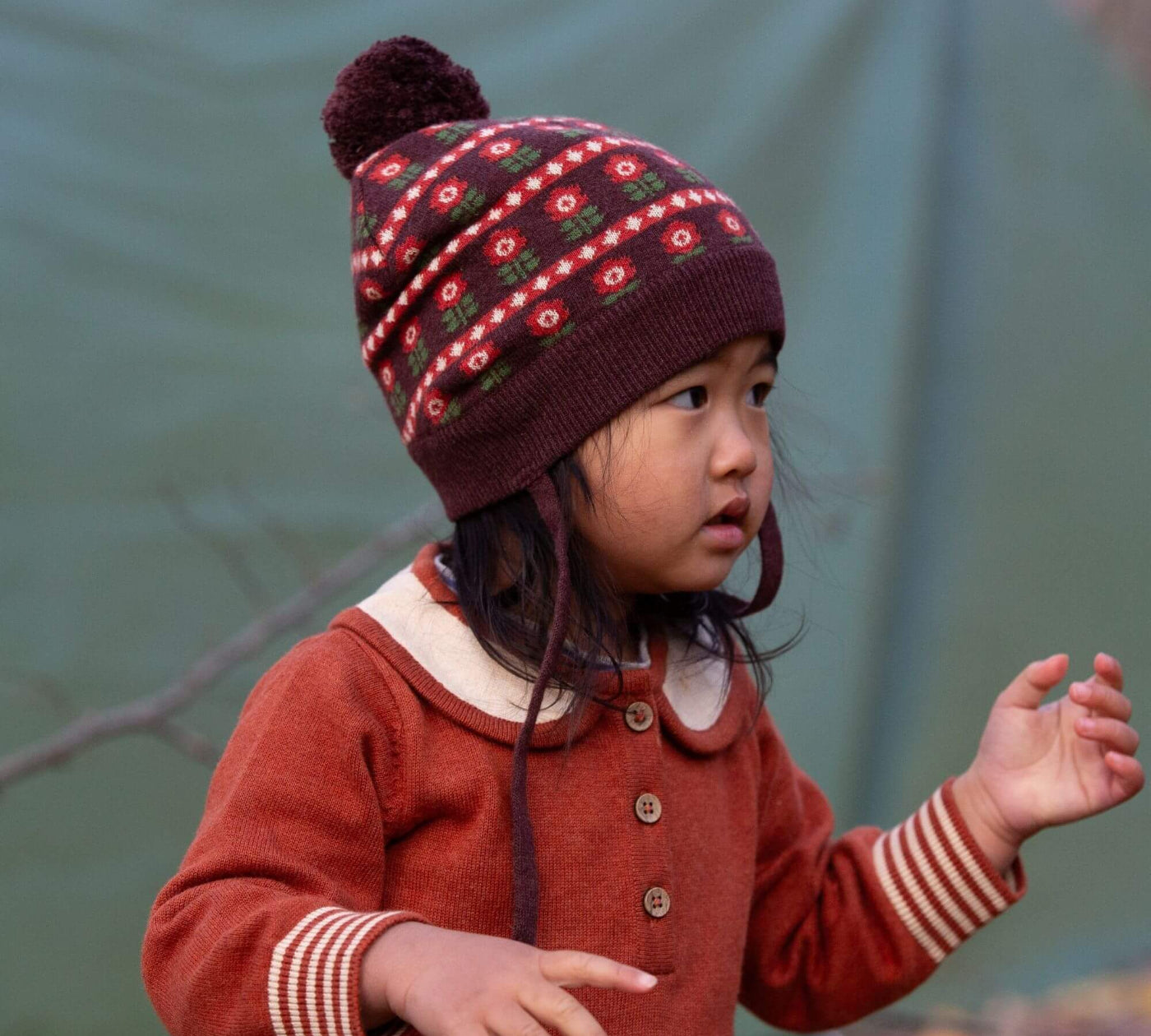 Child wearing a patterned knit hat and ginger knitted dress against a green background