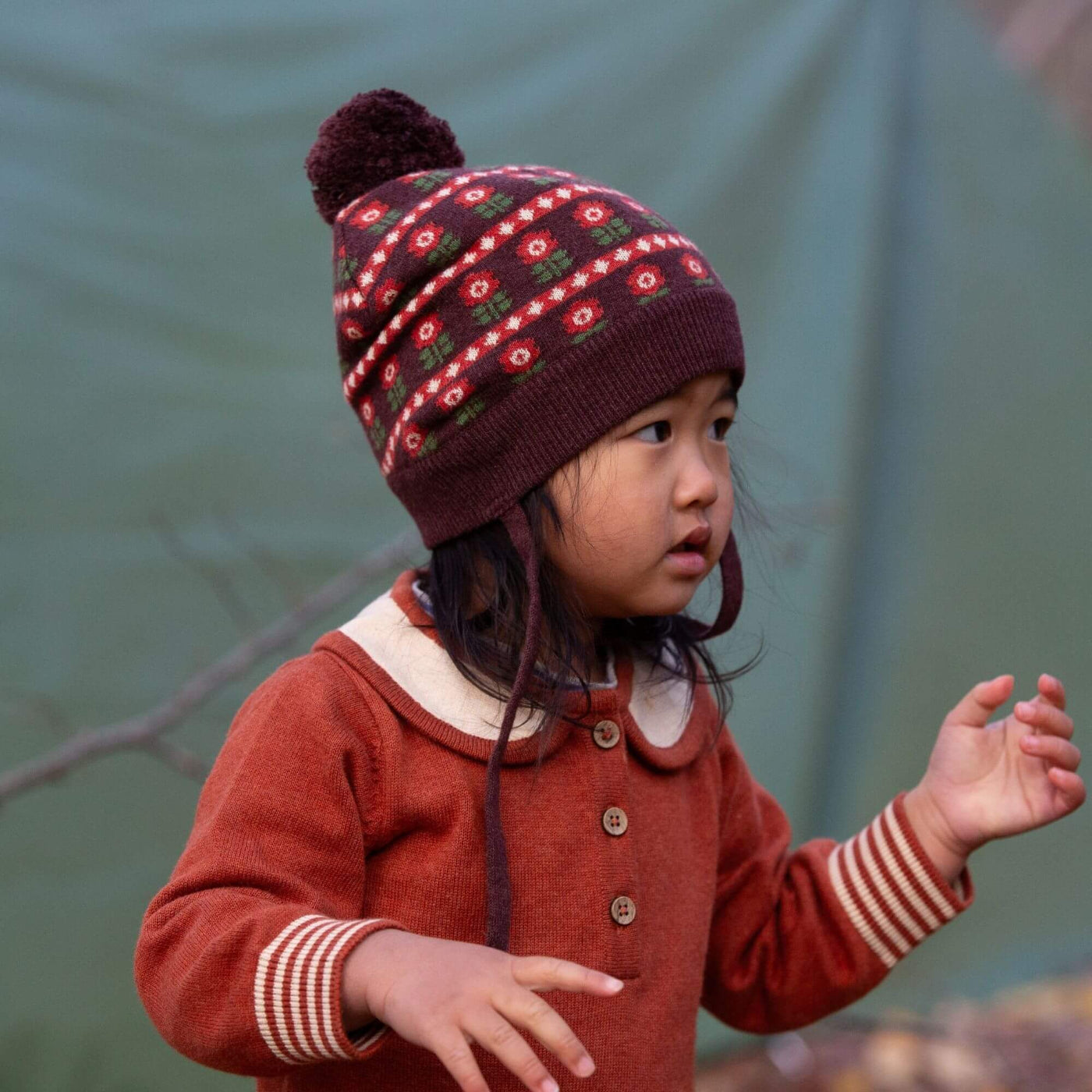Child wearing a patterned knit hat and ginger knitted dress against a green background