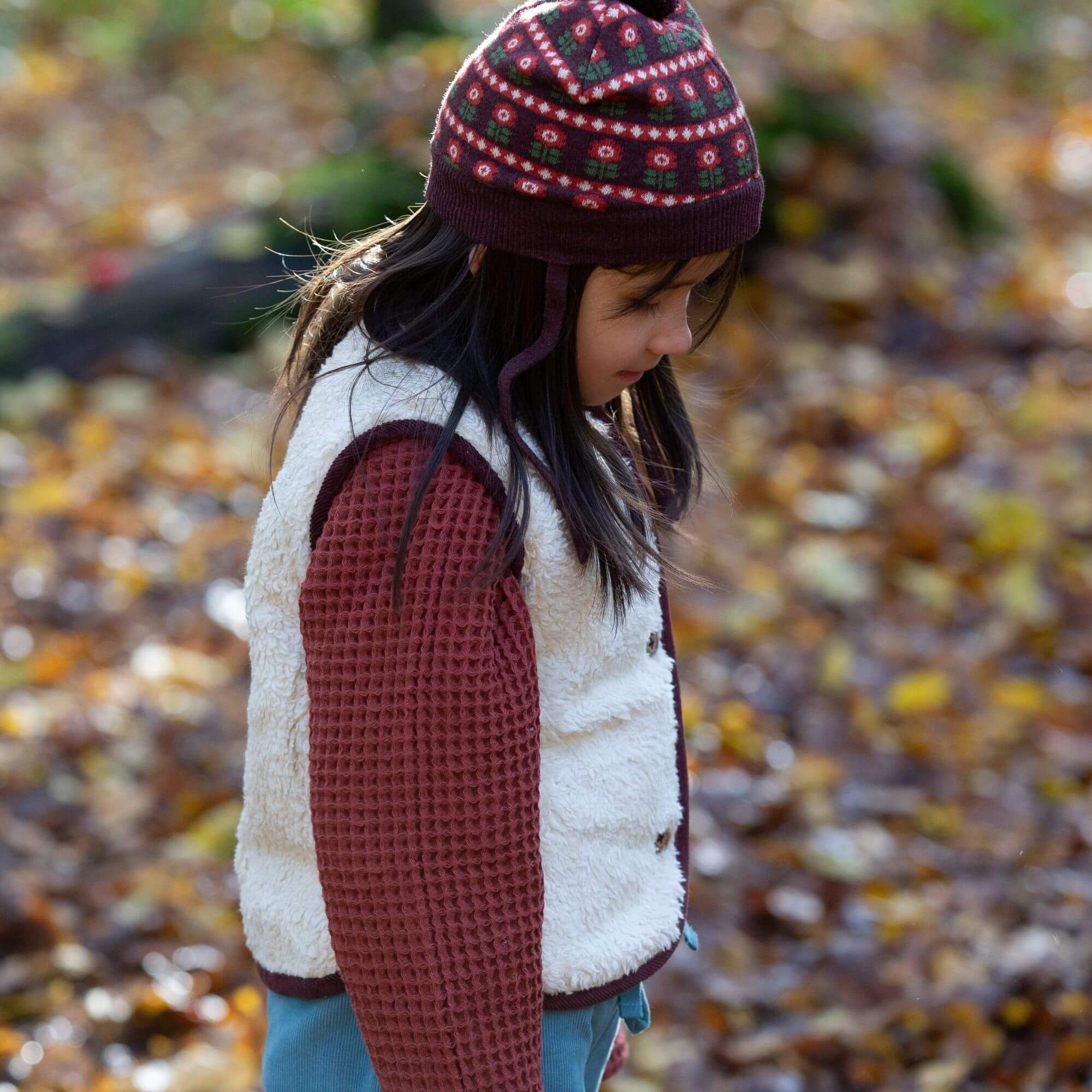 Child wearing a patterned hat and colorful jacket standing in an autumn setting with fallen leaves.