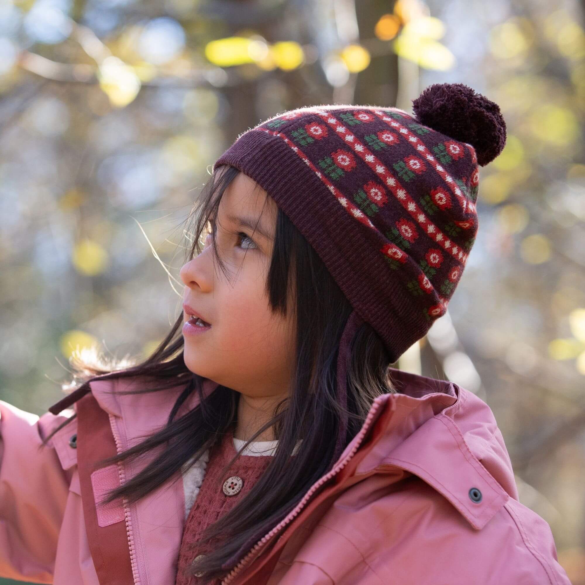 Child wearing a patterned beanie and pink coat in a forest setting